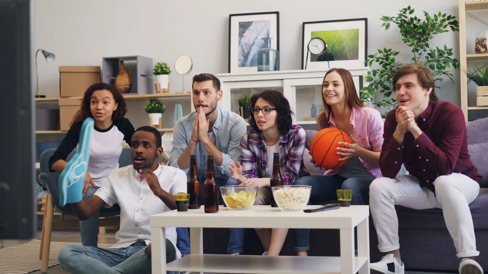Grupo de amigos viendo un partido de básquetbol en la televisión con cervezas y botanas